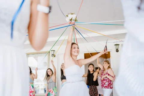 Mujer con vestido blanco sostiene un ramo elevado y unas tijeras mientras varias cintas de colores se extienden hacia otras mujeres.