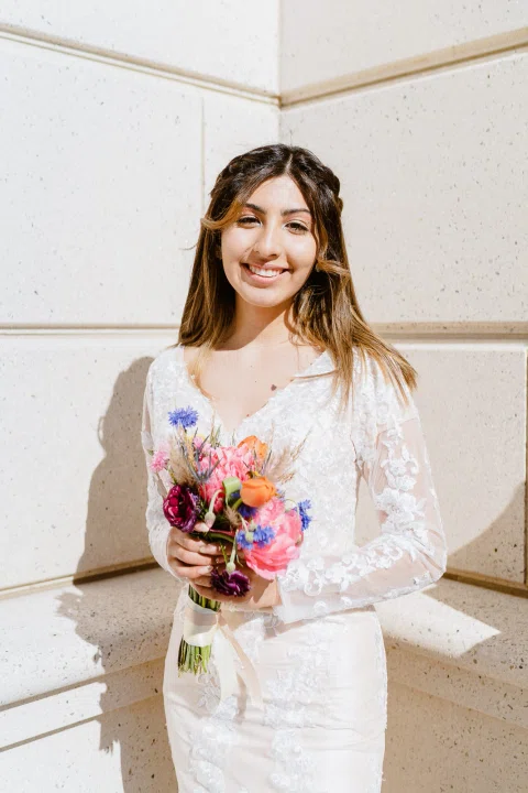 Mujer con vestido blanco posa sola con un ramo de flores coloridas frente a un fondo claro.