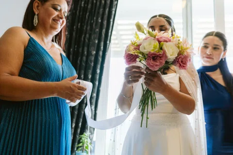 Mujer con vestido blanco sostiene un ramo entre dos mujeres dentro de una habitación.