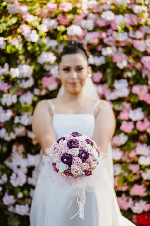 Mujer con vestido blanco posa sola con un ramo frente a abundantes flores rosadas.