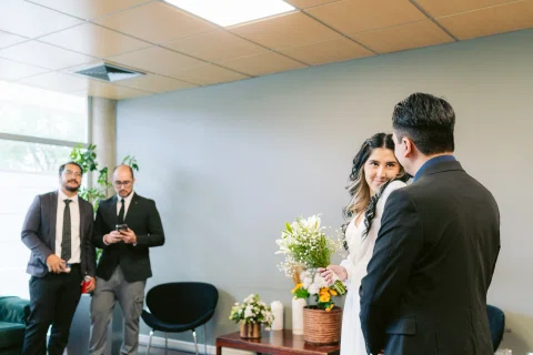Mujer con vestido blanco y ramo permanece frente a un hombre de traje negro, con dos hombres al fondo.