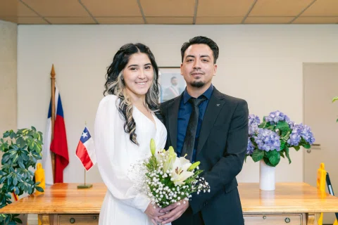 Mujer con vestido blanco y ramo posa junto a un hombre de traje negro frente a banderas y arreglos florales.