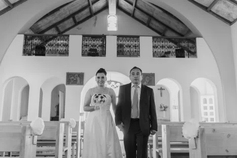 Mujer con vestido blanco y ramo está de pie junto a un hombre con traje oscuro dentro de una iglesia, en blanco y negro.