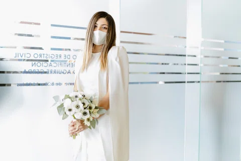 Mujer con vestido blanco, mascarilla y ramo posando frente a un muro de vidrio.