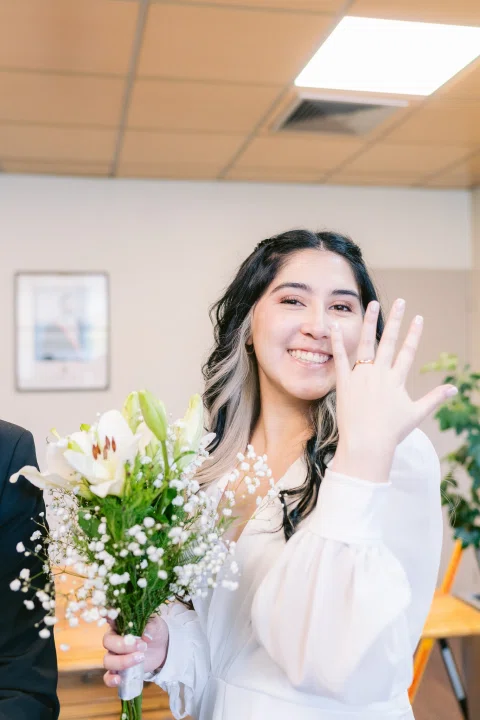 Mujer con vestido blanco y ramo sonríe mientras levanta una mano mostrando un anillo.