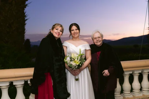 Mujer con vestido blanco y ramo posa entre dos mujeres al aire libre al atardecer.