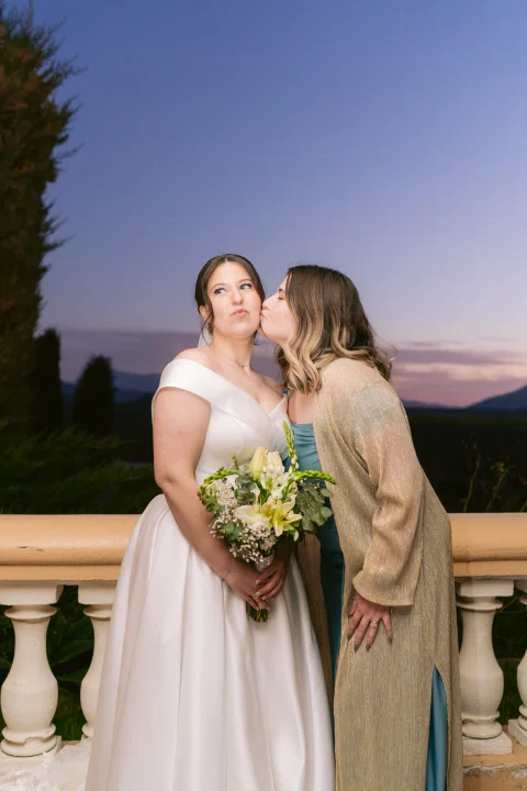 Mujer con vestido blanco y ramo recibe un beso en la mejilla de otra mujer al atardecer.