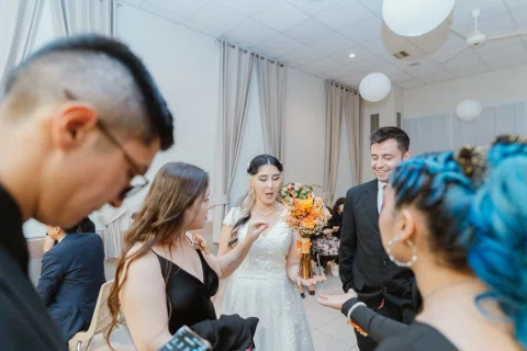 Mujer con vestido blanco sostiene un ramo de flores naranjas mientras varias personas la rodean en un salón.