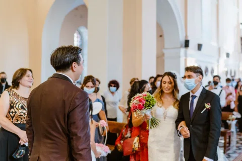 Mujer con vestido blanco y ramo rojo camina por el pasillo de una iglesia mientras varias personas la observan.