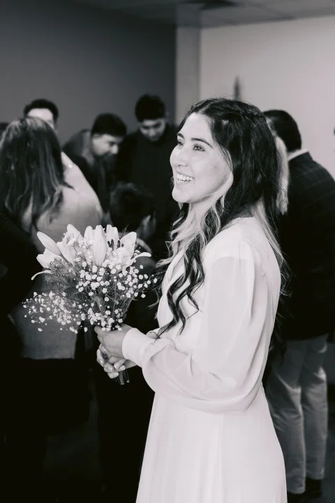 Mujer con vestido blanco y ramo sonríe frente a otra mujer en una fotografía en blanco y negro.