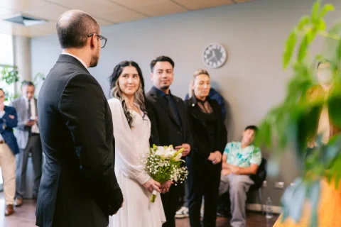 Mujer con vestido blanco y ramo sonríe mientras varias personas permanecen a su alrededor.
