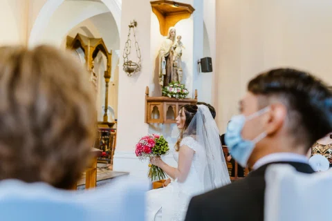 Mujer con vestido blanco y ramo vista entre varias personas sentadas dentro de una iglesia.