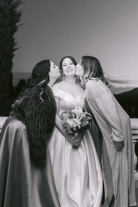 Mujer con vestido blanco recibe besos en ambas mejillas de dos mujeres, en blanco y negro.