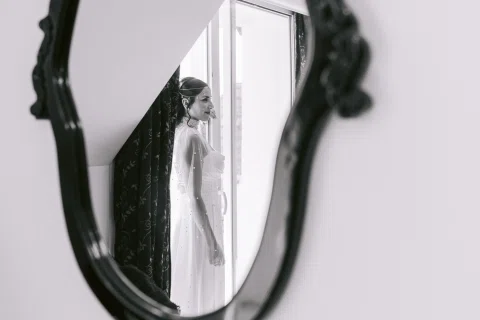 Mujer con vestido blanco aparece reflejada en un espejo junto a un ventanal.