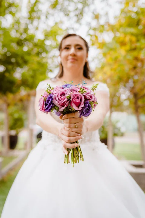 Mujer con vestido blanco posa sola con un ramo de flores rosadas en el jardín.