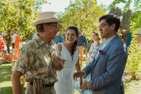 Mujer con vestido blanco ríe junto a dos hombres en el jardín.