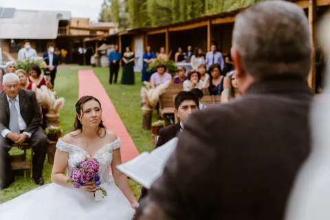 Mujer con vestido blanco está sentada con un ramo mientras un hombre sostiene un documento en primer plano.