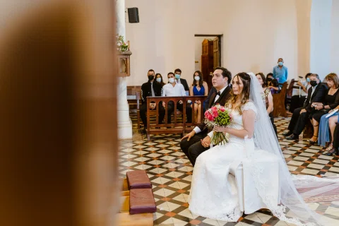 Mujer con vestido blanco sentada sosteniendo un ramo de flores rojas dentro de una iglesia.