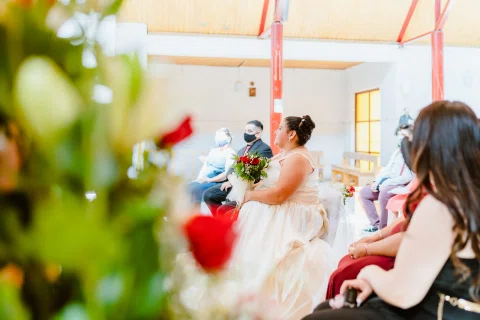Mujer con vestido blanco sentada con un ramo, con flores rojas desenfocadas en primer plano.