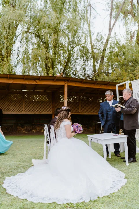 Mujer con vestido blanco está sentada de espaldas con el vestido extendido sobre el césped frente a dos hombres.