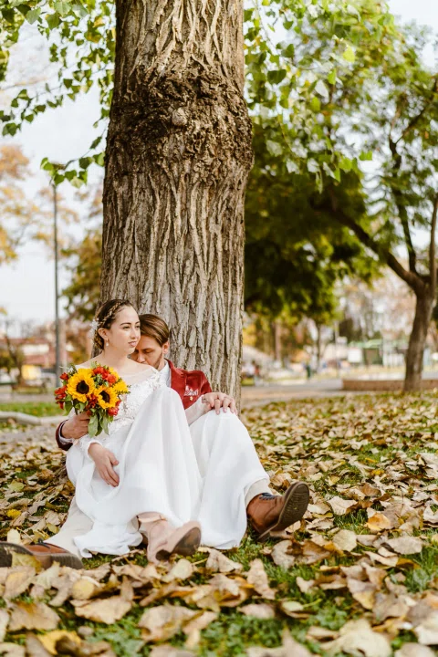 Mujer con vestido blanco está sentada al pie de un árbol junto a un hombre con chaqueta roja; sostiene un ramo de flores.