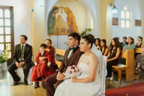 Mujer con vestido blanco está sentada con un ramo rosado mientras varias personas permanecen detrás de ella.