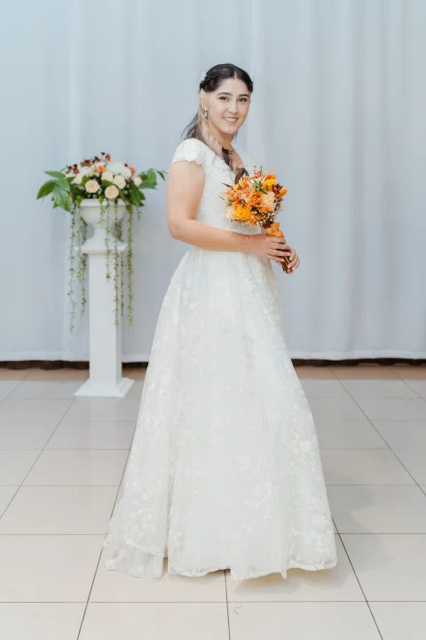 Mujer con vestido blanco posa sola de cuerpo entero sosteniendo un ramo de flores naranjas.