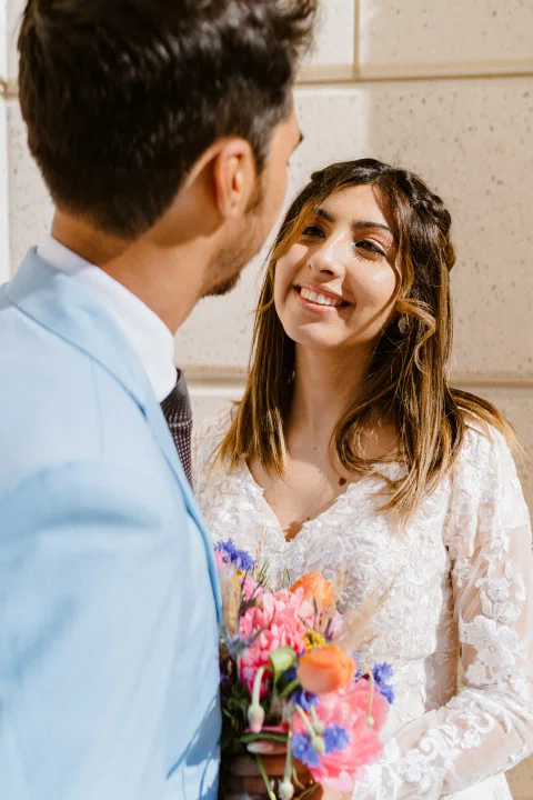 Mujer con vestido blanco sonríe frente a un hombre con traje celeste mientras sostiene un ramo colorido.