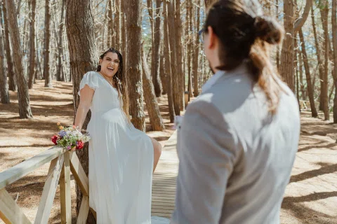 Mujer con vestido blanco sonríe frente a un hombre con traje gris mientras sostiene un ramo entre árboles.