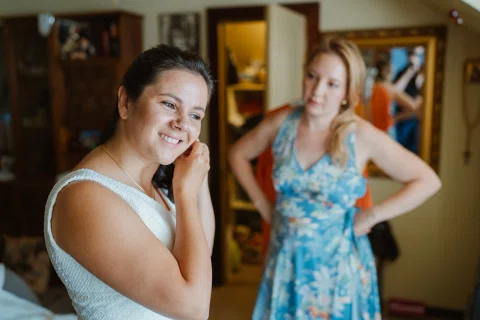 Mujer con vestido blanco sonríe mientras se toca una oreja; otra mujer con vestido azul estampado está detrás.