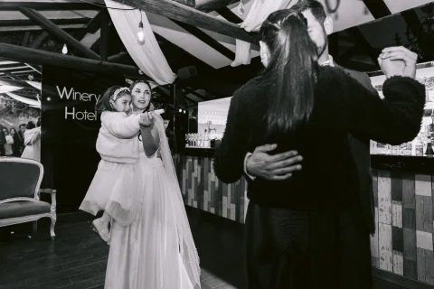 Mujer con vestido blanco sostiene en brazos a una niña mientras un hombre y una mujer bailan en primer plano, en una fotografía en blanco y negro.