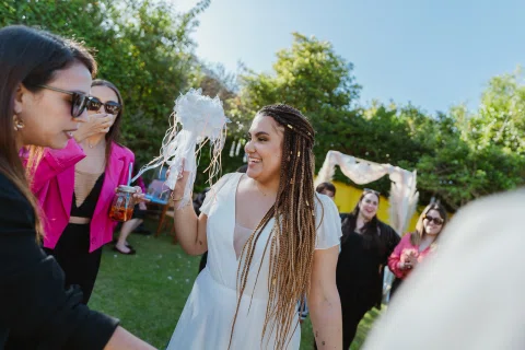 Mujer con vestido blanco sostiene un ramo blanco frente a varias mujeres reunidas en el jardín.