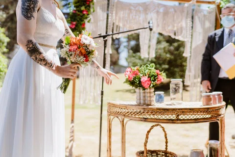 Mujer con vestido blanco sostiene un ramo junto a una mesa con flores.