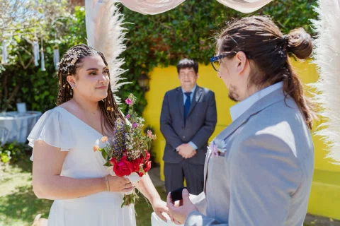 Mujer con vestido blanco sostiene un ramo mientras mira a un hombre con traje gris bajo el arco.