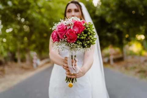 Mujer con vestido blanco sostiene un ramo rojo frente a su rostro en un retrato exterior.