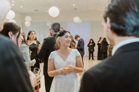 Mujer con vestido blanco sostiene un vaso mientras conversa con una persona vista de espaldas en un salón.