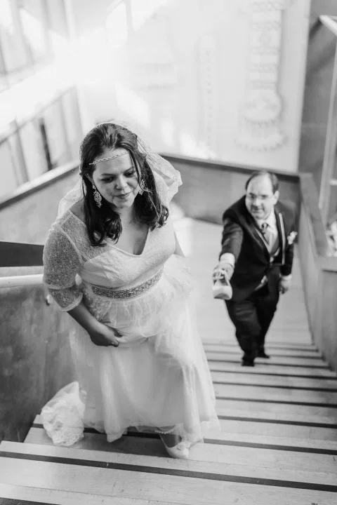 Mujer con vestido blanco sube una escalera mientras un hombre con traje oscuro avanza detrás de ella, en una fotografía en blanco y negro.