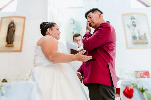 Mujer con vestido blanco toca el rostro de un hombre con chaqueta burdeos frente al altar.