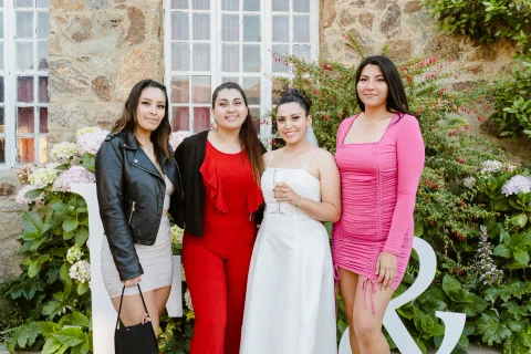 Mujer con vestido blanco posa con otras tres mujeres frente a un muro de piedra y vegetación.