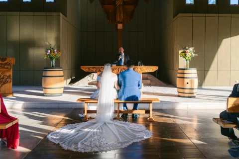 Mujer con vestido blanco y velo y hombre con traje azul están sentados de espaldas frente al altar.
