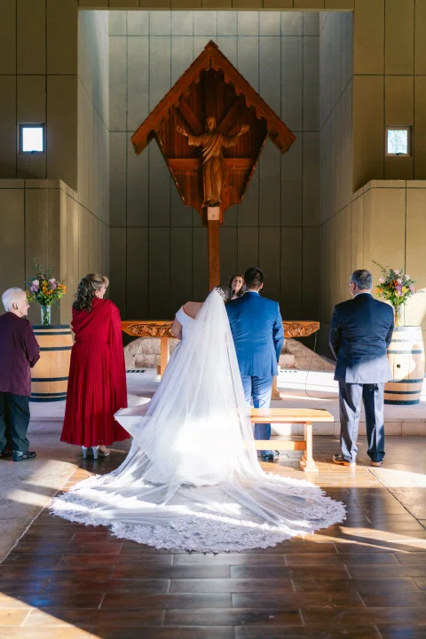 Mujer con vestido blanco y velo permanece junto a un hombre con traje azul frente al altar, vistos desde atrás.