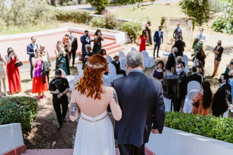 Mujer con vestido blanco y un hombre con mascarilla permanecen frente a un grupo de personas en un jardín.