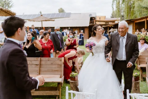 Mujer con vestido blanco y un hombre con traje oscuro avanzan por la alfombra roja entre personas sentadas.