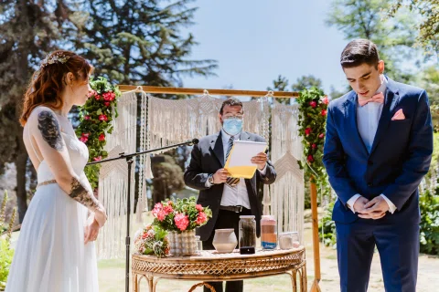 Mujer con vestido blanco y hombre con traje azul permanecen junto a una mesa con flores y pequeños frascos mientras otra persona sostiene documentos.