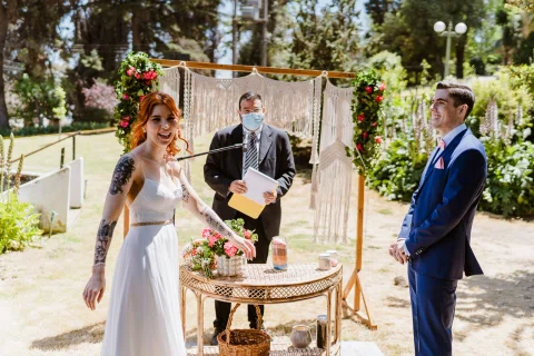Mujer con vestido blanco y hombre con traje azul permanecen junto a una mesa redonda con flores mientras otra persona sostiene documentos.