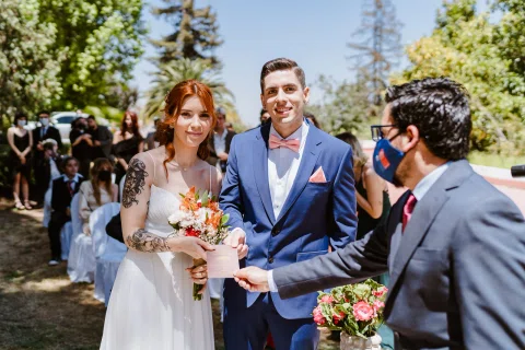 Mujer con vestido blanco y hombre con traje azul sonríen en un jardín mientras varias personas permanecen detrás.