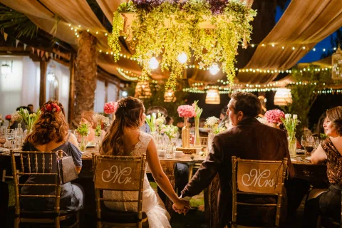 Mujer con vestido blanco y hombre con traje burdeos permanecen sentados en una mesa tomados de la mano, con otras personas alrededor bajo luces cálidas.