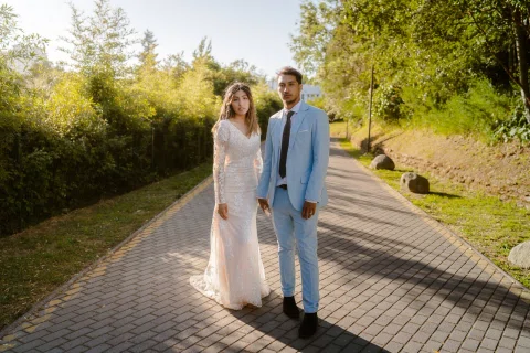 Mujer con vestido blanco y hombre con traje celeste permanecen de pie sobre un sendero pavimentado entre árboles.