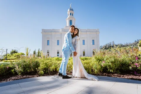 Mujer con vestido blanco y hombre con traje celeste se besan en el jardín frente al edificio blanco.