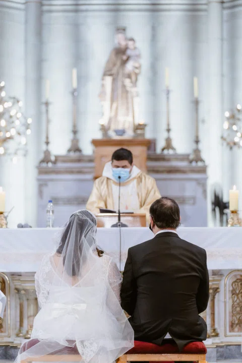 Mujer con vestido blanco y hombre con traje oscuro permanecen arrodillados frente al altar de una iglesia.
